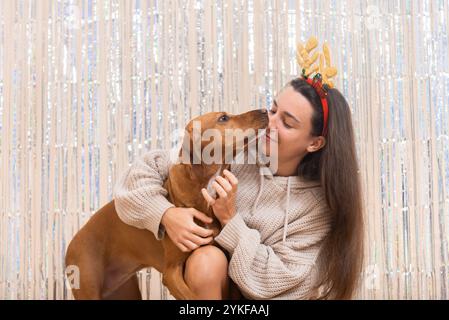 Eine Frau mit weihnachtlichem Rentiergeweih hält liebevoll und tauscht sich mit ihrem braunen Hund in einer gemütlichen Innenumgebung aus Stockfoto