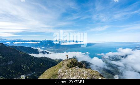 Eine Person, die auf dem zerklüfteten Gipfel des Dent de Jaman steht, mit triumphalen Armen unten, ein Wolkenmeer bedeckt die Landschaft, während die blauen Schweizer Stockfoto