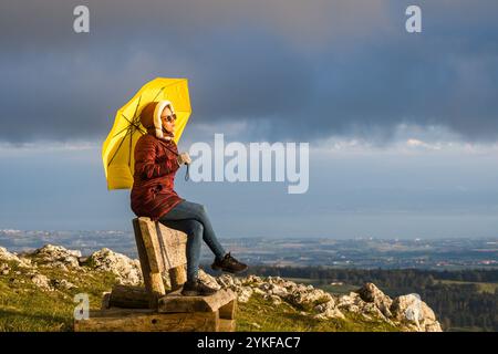 Seitenansicht einer Frau, die den atemberaubenden Sonnenuntergang über dem Schweizer Jura von Dent de Vaulion aus genießt, auf einer Holzbank mit einem leuchtend gelben Regenschirm, Stockfoto