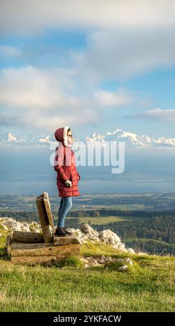 Seitenansicht einer Frau in einer roten Jacke steht auf einer Holzbank mit Blick auf den malerischen Dent de Vaulion während des Sonnenuntergangs. Mit dem schneebedeckten Schweizer Jura Mo Stockfoto