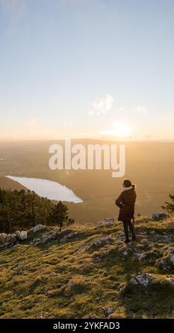 Rückansicht einer nicht erkennbaren Person, die auf einem Berg im Dent de Vaulion im Schweizer Jura steht und einen atemberaubenden Sonnenuntergang über einem malerischen Tal bestaunt Stockfoto