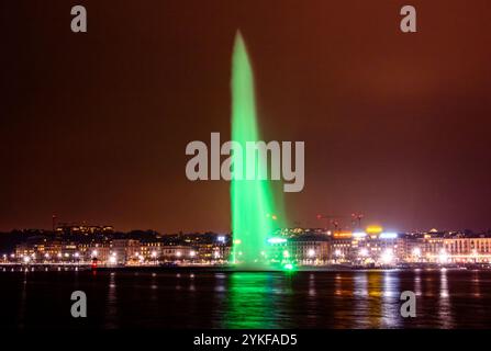 Ein atemberaubender Blick bei Nacht auf einen hellgrünen Brunnen, der hoch über dem Genfer See schwingt, mit den Lichtern der Stadt Genf im Hintergrund Stockfoto