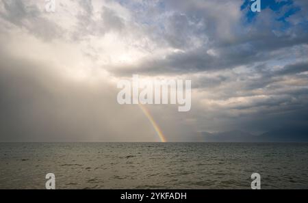 Atemberaubende Regenbogenbögen über dem nebeligen Wasser des Genfer Sees in Morges, Schweiz, werden von einem Hintergrund aus bewölktem Himmel und fernen Bergen abgehoben Stockfoto