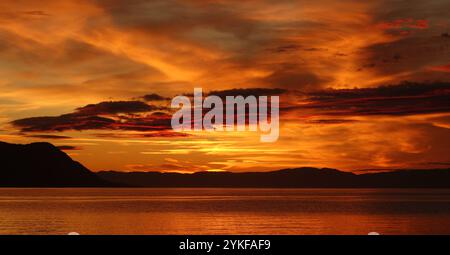 Ein atemberaubender Sonnenuntergang färbt den Himmel in leuchtenden Orange- und Rottönen und reflektiert auf dem ruhigen Wasser die Silhouetten der Berge verleihen Tiefe, während sie dramatisch sind Stockfoto