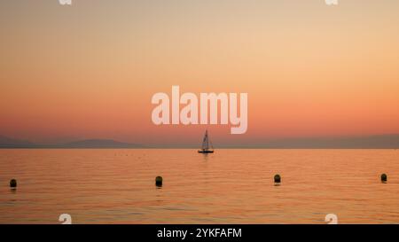 Ein einsames Segelboot driftet auf einem ruhigen Meer unter dem sanften Glanz eines Pfirsichs und rosa Sonnenuntergangs sanfte Wellen und ferne Hügel schaffen eine ruhige Meereslandschaft, evo Stockfoto