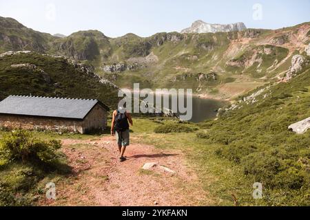 Ein Wanderer geht einen unbefestigten Pfad hinunter zu einem ruhigen Bergsee, der von zerklüfteten Gipfeln in Asturien, Spanien, umgeben ist, was ein perfektes Outdoor-Abenteuer darstellt Stockfoto