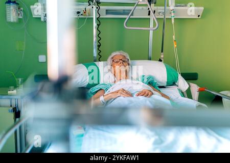 Ältere Frau, die in einem Krankenhausbett liegt, in einen grünen Krankenhausmantel gewickelt, ist der Raum mit medizinischen Geräten ausgestattet und hat ein ruhiges, grünes Ambiente, Empha Stockfoto