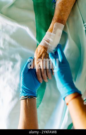 Ein medizinisches Fachpersonal mit blauen Handschuhen hält sanft die Hand einer älteren Frau, die in einem Krankenhausbett ruht. Die Szene vermittelt Empathie, Unterstützung, und Stockfoto