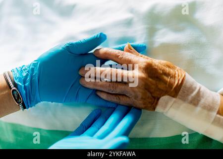 Eine ältere Frau in einem Krankenhausbett erhält Komfort von einer Krankenschwester mit Handschuhen die Krankenschwester hält sanft die Hand des Patienten und veranschaulicht Mitgefühl und c Stockfoto