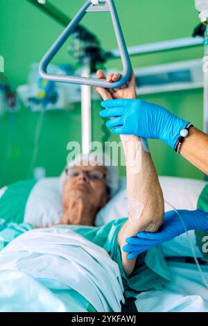 Eine ältere Frau liegt in einem Krankenhausbett, wobei ihr Arm von einem medizinischen Fachpersonal mit Schutzhandschuhen sanft gestützt wird Stockfoto