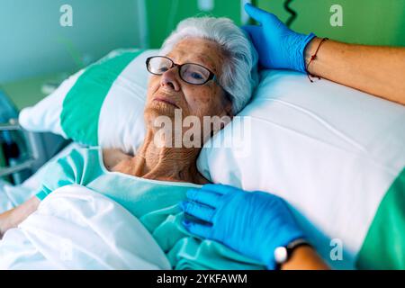 Eine ältere Frau liegt in einem Krankenhausbett mit der Hand einer Pflegeperson sanft auf ihre Schulter gelegt Sie wirkt ruhig, trägt eine Brille, während die surr Stockfoto