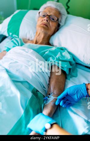 Eine ältere Frau mit grauen Haaren und Brille liegt in einem Krankenhausbett und erhält medizinische Behandlung Ein medizinisches Fachpersonal in blauen Handschuhen stellt sie I ein Stockfoto