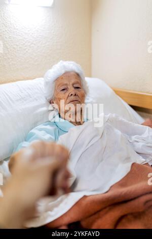 Eine ältere Frau liegt im Bett und hält die Hände mit jemandem außerhalb des Rahmens. Die Szene vermittelt ein Gefühl von Sorgfalt und Gesellschaft und hebt Themen von älteren Menschen hervor Stockfoto