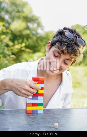 Eine fröhliche, nicht-binäre Person mit kurzen lockigen Haaren baut einen bunten Turm aus Holzblöcken auf einem Tisch im Freien, der fokussiert und fröhlich in einem nat aussieht Stockfoto