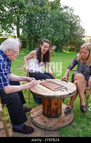 Eine kaukasische Familie spielt an einem sonnigen Tag gerne ein Brettspiel im Freien. Das Bild zeigt einen älteren Mann, eine junge Frau und eine Frau mittleren Alters im Sitzen Stockfoto