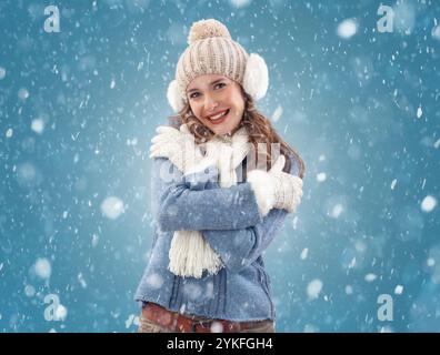 Fröhliche Frau, die die Schönheit eines Winterschneefalls umschmeichelt. Sie lächelt über den fallenden Schnee während eines weißen Weihnachtsfestes. Tragen einen kuscheligen blauen Pullover, Bommel... Stockfoto
