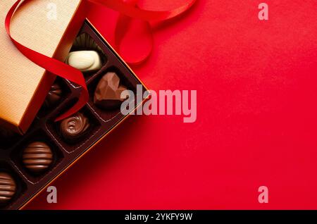 Verschiedene Schokoladenbonbons in einer goldenen Schachtel und einem roten Band, auf rotem Hintergrund mit Kopierraum. Stockfoto