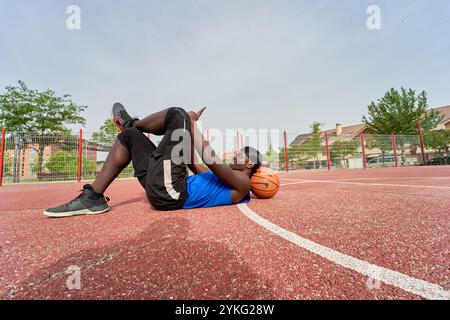 Ein Mann liegt auf dem Boden mit einem Basketball im Kopf mit ihrem Smartphone. Er trägt ein blaues Hemd und schwarze Shorts Stockfoto