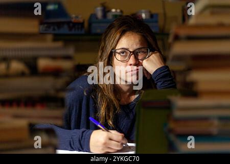 Gestresste junge Frau mit Brille studiert für Prüfungen, umgeben von Büchern in der Nacht Stockfoto