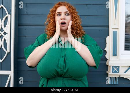 Überraschte Plus Size Frau mit lockigem rotem Haar in grüner Kleidung gegen bunten blauen Wand Hintergrund im Freien, Modell mit schockiertem Gesichtsausdruck, Stockfoto