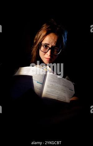 Gestresste junge Frau mit Brille studiert für Prüfungen, umgeben von Büchern in der Nacht Stockfoto