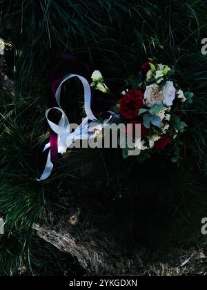 Ein eleganter Brautstrauß mit roten und weißen Rosen mit Grün, verziert mit fließenden Bändern, ruht auf üppigem grünem Gras und schafft eine markante Mischung Stockfoto