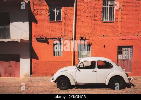 Guanajuato, Mexiko - 4. Februar 2024: Ein klassischer weißer Volkswagen-Käfer oder Bug parkt an einer leuchtend roten Wand unter der mexikanischen Sonne und fängt ein Stockfoto