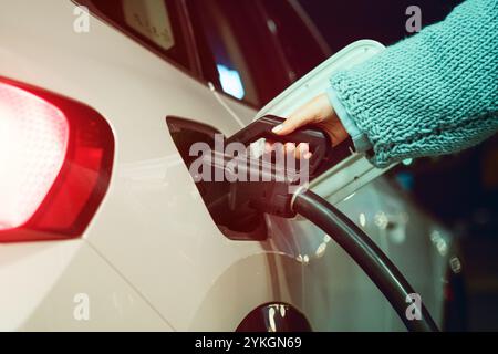 Eine Nahaufnahme einer Frauenhand, die ein Ladekabel an einem weißen Elektrofahrzeug an einer Ladestation anschließt und dabei saubere Energie und moderne Energie betont Stockfoto
