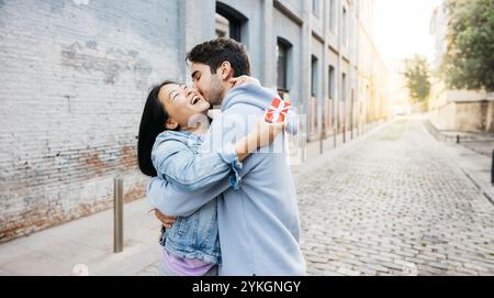 Glückliche asiatische Frau, die ihren Freund umschließt, nachdem sie ein Geschenk zum Valentinstag erhalten hat. Verliebtes Paar auf der Straße, um den Valentinstag zu feiern Stockfoto