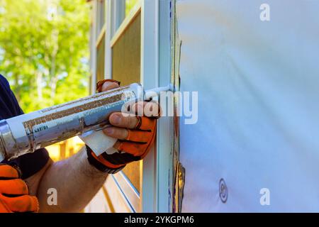 Bei Wohnbauprojekten trägt der Arbeiter mit Schutzhandschuhen sorgfältig Dichtungen an den Kanten des Fensterrahmens auf, um eine dichte Abdichtung gegen Luftlecks zu gewährleisten. Stockfoto