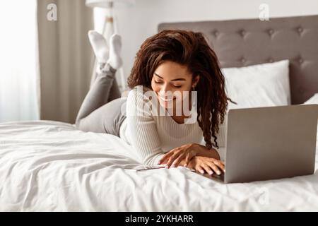 Positive schwarze Frau, die im Bett liegt und eine Textnachricht liest Stockfoto