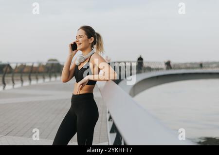 Eine Joggerin hält an ihrem Telefon am Fluss an, trägt Trainingsausrüstung und Kopfhörer und genießt die warme Sonne und die ruhige Atmosphäre Stockfoto