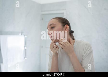 Gen z Frau, die Pickel aufs Kinn wirft, steht vor einem Spiegel im Badezimmer. Konzept der Aknebehandlung, Hautpflege, Akne Erwachsener Stockfoto