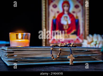 Die Zeit des Gebets. Hölzerner Rosenkranz auf einem alten Gebetsbuch. Ikone der Mutter Gottes und Jesu. Beleuchtete Kerze. Stockfoto