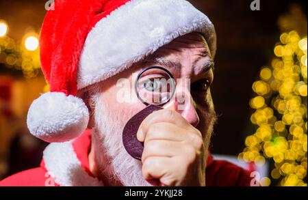 Weihnachtsmann, der durch die Lupe schaut. Weihnachten oder Neujahr. Bärtiger Mann mit Weihnachtsmütze mit Lupe, Nahaufnahme Porträt. Santa man Toilette Stockfoto