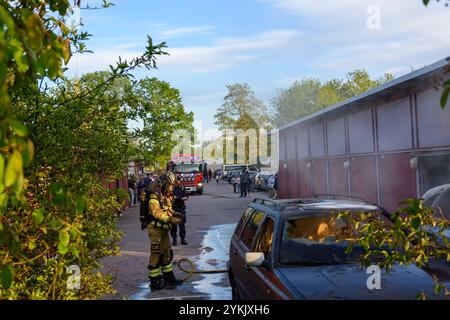 Notfallhelfer arbeiten fleißig daran, einen Autobrand neben einer Garage in einer Wohngegend zu kontrollieren. Als Zuschauer bläst Rauch aus dem Fahrzeug Stockfoto