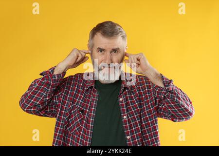 Senior-Mann, der seine Ohren mit Fingern auf orangefarbenem Hintergrund bedeckt Stockfoto