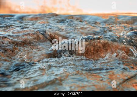 Wasser stürzt über glatte Felsen und erfasst die dynamische Bewegung eines fließenden Baches. Das Sonnenlicht reflektiert das Wasser und verleiht ihm ein warmes und lebendiges Leuchten. Stockfoto