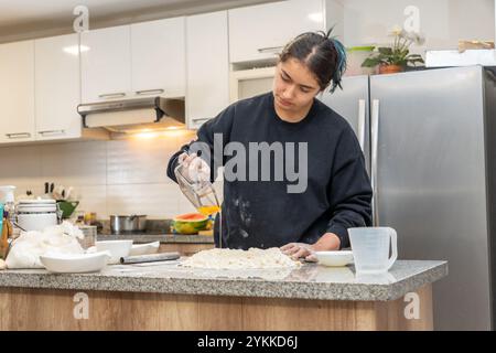 Die junge Latina mit schwarzen Haaren, die gefesselt sind, legt rohe Eier in Mehl, um Teig auf die Küchenzeile in ihrem Haus zu machen Stockfoto