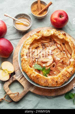 Eine frisch gebackene Apfeltorte sitzt auf einem rustikalen Holzbrett, garniert mit Mandeln und Minze, begleitet von roten Äpfeln und einer kleinen Schüssel Honig, Inviti Stockfoto