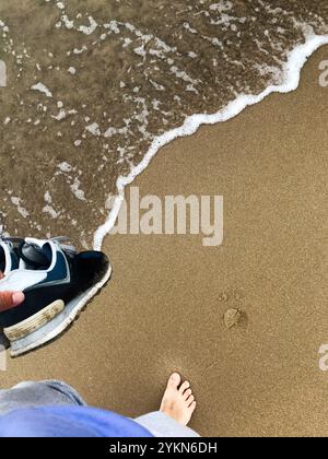 Eine Person genießt einen friedlichen Barfußspaziergang an einem Sandstrand mit Sneakers in der Hand, während sanfte Wellen an Land strömen und das Wesen der Entspannung einfangen Stockfoto