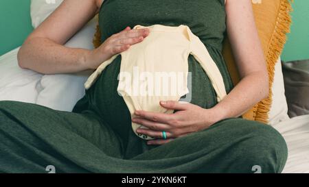 Die werdende Mutter sitzt bequem mit Babykleidung auf dem Bauch Stockfoto