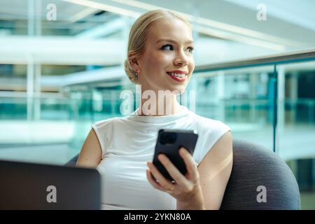 Eine selbstbewusste Buchhalterin sitzt in einem modernen Büroumfeld, hält ein Smartphone und lächelt und unterstreicht ihr Leistungsgefühl Stockfoto