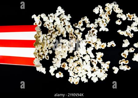 Blick von oben auf einen rot-weißen Pappeimer, der flach mit streuendem Popcorn auf schwarzem Hintergrund liegt, Konzept für Essen und Kinounterhaltung Stockfoto