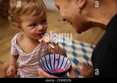 Ein niedliches Kleinkind isst gerne aus einer gestreiften Schüssel, während es einen fröhlichen Moment mit einem Familienmitglied teilt. Das Bild fängt Wärme und Zuneigung in einem Casua ein Stockfoto