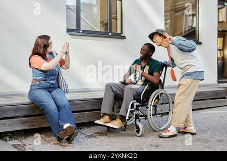 Freimütige Szene in voller Länge, in der ein junger afroamerikanischer Mann mit Behinderung Fotos mit Freunden im Freien macht und gemeinsam Spaß hat Stockfoto