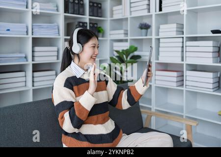 Eine junge Frau nimmt mit ihrem Tablet freudig an einem Videoanruf Teil und präsentiert ein komfortables Home Office-Setup mit minimalistischer und moderner Ästhetik. Stockfoto