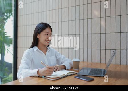 Eine junge Frau sitzt an einem stilvollen Café-Tisch, arbeitet an einem Laptop und schreibt Notizen in ihr Notizbuch, während sie einen warmen Kaffee genießt. Stockfoto