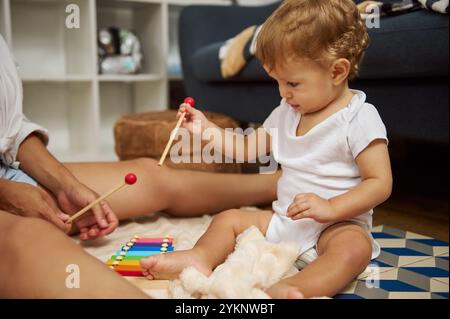 Ein junges Kleinkind spielt fröhlich mit einem farbenfrohen Xylophone auf dem Boden, umgeben von einer warmen, gemütlichen Wohnkultur. Die Szene fängt einen Moment des Lernens ein Stockfoto