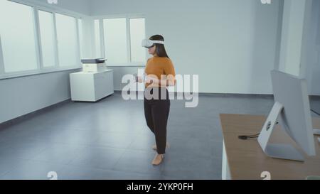 Kreative asiatische Designerin, die ein Augmented-Reality-Headset trägt, nutzt Virtual Interior Design Software und Controller, um einen Büroraum mit 3D-Möbeln und -Geräten zu bauen. Futuristische VFX-Animation. Stockfoto